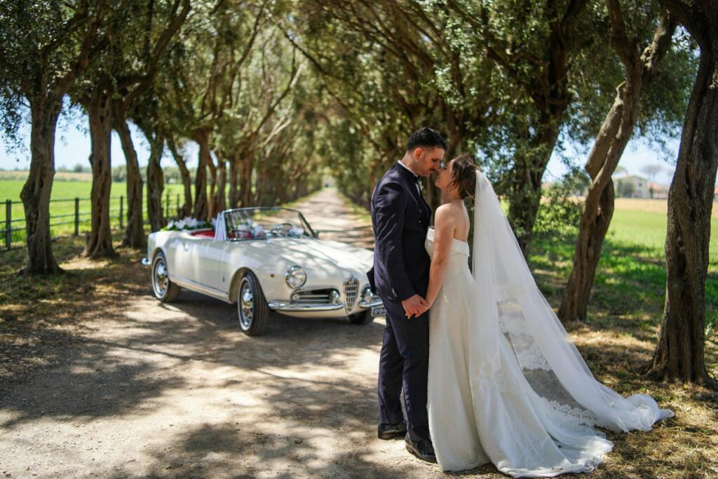 Bride and groom beside a vintage car on a tree-lined road